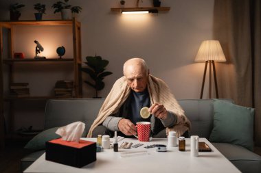 Battaniyeye sarılmış yaşlı bir adam kırmızı bir benekli bardakta limon çayı hazırlıyor. İlaçlar onun önündeki masaya yayıldı. Arka planda metin için alanı olan otantik ev atmosferi.