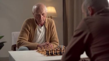 Yaşlı bir adam ve arkadaşı yoğun bir şekilde sıcak, aydınlık bir oturma odasında satranç oynamaya odaklanır..