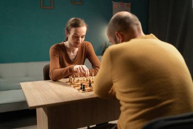 Uzun saçlı genç bir adam ve sarı kazaklı kel bir adam rahat bir ev ortamında satranç oynuyorlar. Tahta satranç tahtası ahşap bir masanın üzerine konur.