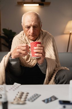 Battaniyeye sarılı yaşlı bir adam elinde bir fincan çay tutuyor. Evde, çeşitli ilaçlarla çevrili, sağlığını kontrol etme ve hastalıktan kurtulma çabalarını gösteriyor..