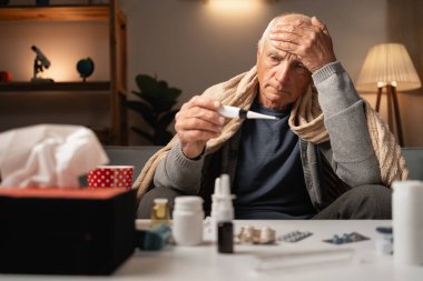 Yüksek ateşi olan yaşlı bir adam, evdeyken termometresini dikkatlice kontrol ediyor. Etrafı çeşitli ilaçlarla çevrili bir battaniyeye sarılmış ve kendini pek iyi hissetmiyor ve endişeli görünüyor..