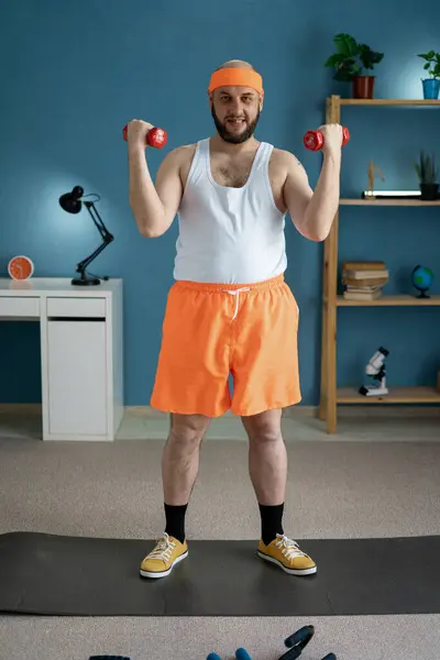 Stock image Funny middle-aged man in orange shorts and headband lifting red dumbbells in his home gym with a smile.