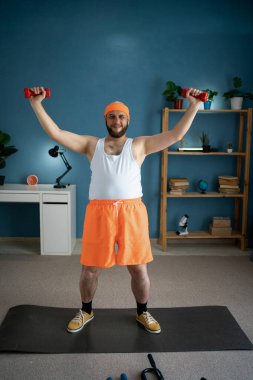 Evde yoga minderinin üzerinde duran orta yaşlı bir adam vücudunun üst kısmını güçlendirmek için halterleri kaldırıyor. O, ev fitness rutini ile kendinden emin görünüyor. Ev sağlığı ve sağlıklı yaşam tarzı kavramı.