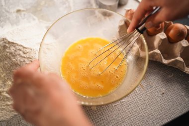 Masada un ve yumurta olan bir kasede yumurta çırpan ellerin yakın çekimi. Yemek pişirme, yemek hazırlama ve pişirme kavramı. Üst görünüm