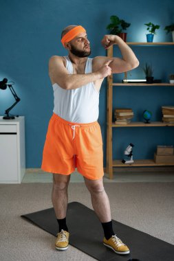 Kendine güveni tam bir adam evde çalışırken kaslarını esnetiyor ve kas gelişimini gururla gösteriyor. Turuncu şort, saç bandı, sağlıklı yaşam tarzı ve vücut dönüşümü konsepti.