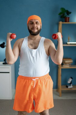 Kırmızı dambılları kaldıran bir adam spor salonunda kol çalışmasına odaklanmış. Turuncu bir saç bandı, beyaz bir atlet ve turuncu bir şort giyerek kaslarını güçlendirirken kendine güvenerek ayakta duruyor..