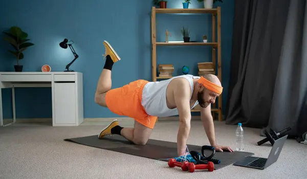 stock image Man exercising at home while watching an online workout video on his laptop. He is lifting his leg on a yoga mat in a room filled with fitness equipment including dumbbells, a jump rope, and a water bottle.