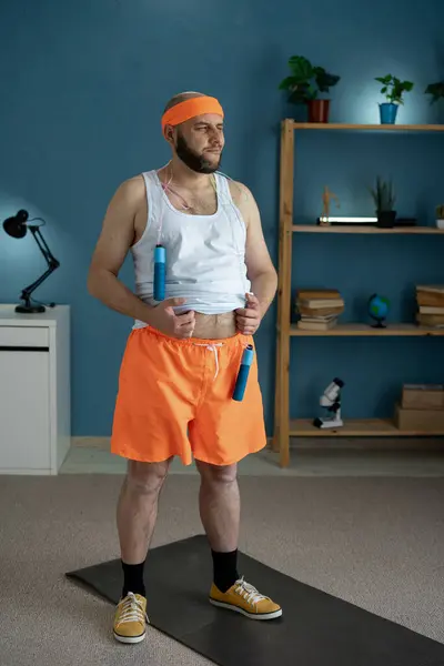 stock image Man with excess weight is preparing for his home fitness session, adjusting his clothes and thoughtfully considering a jump rope workout. concept of home fitness and weight loss.