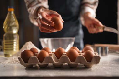 Bir kadının eli yumurta kartonundan yumurta topluyor, yumurta ve karton üzerine odaklanmış bir şekilde. Yemek malzemeleri masanın üzerine dizilmiş..
