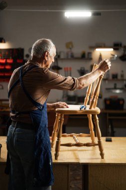 Mobilya onarımı, DIY ve ev iyileştirme konsepti. Atölyedeki yaşlı adam restorasyona başlamadan önce eski ahşap sandalyeyi teftiş ediyor..