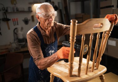 Atölyedeki yaşlı bir adam ahşap bir sandalyeden eski verniği çıkarmak için özel eklenmiş matkap kullanıyor. Koruyucu gözlük ve turuncu eldiven takıyor. konsept mobilya restorasyonu