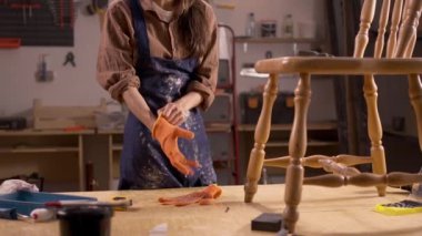 Atmosferik atölyede genç bir kadın eski bir ahşap sandalyeyi restore etmeye hazırlanıyor, iş eldiveni takıyor. Cihazlar ve malzemeler etrafa saçılmış durumda. Kırsal alanda görevine odaklanıyor. Yüzü olmayan