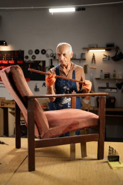Turuncu eldivenli ve mavi önlüklü yaşlı bir adamın portresi atölyede bir sandalyenin ahşap çerçevesini inceliyor. O, sandalyeyi restorasyon için hazırlıyor. Boşluğu kopyala.