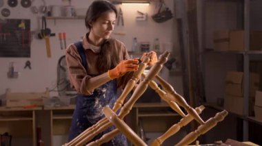 Atmosferik atölyesinde duran beyaz önlüklü kadın zımpara kağıdıyla eski bir sandalyeyi zımparalıyor. Bir parça zımpara kağıdıyla mobilyaları zımparalama işlemi.