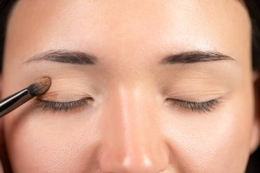 Makyaj fırçası göz kapağına nazikçe sürerken, kapalı gözlü bir kadının detaylı görüntüsü. Titiz makyaj uygulaması, cildinin pürüzsüz dokusunu vurguluyor.