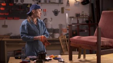 Bir ev atölyesinde, bir kadın eski ahşap bir sandalyeyi onarmaya başlamadan önce eldiven giyer. Arka planda çeşitli araçlar ve ekipmanlar görülebilir, mobilya yenileme projesi.