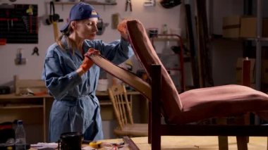 Bir ev atölyesinde, bir kadın eski ahşap bir sandalyenin arkasını kaldırıyor. Çalışma alanı çeşitli araç-gereç ve mobilya restorasyonu süreçleriyle doludur..