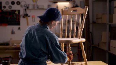 Bir ev atölyesinde, bir kadın onarmak ve restore etmek için eski bir ahşap sandalyeyi inceler. Çalışma alanında çeşitli araç ve gereçler, mobilya restorasyonu süreci sergileniyor.