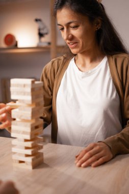 Evinde tahta blok oyunu oynayan gülümseyen kadın bir kule inşa etmek için blokları dikkatlice yerleştiriyor ve dengeliyor. Eğlenceli ve rahatlatıcı bir aktivite, oyuna odaklanma ve kaliteli zaman geçirme.