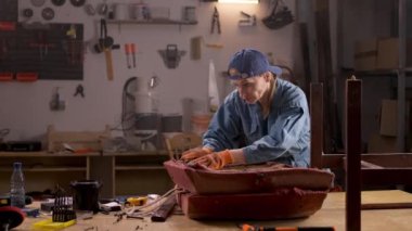 Bir ev atölyesinde, bir kadın eski kumaşı ahşap bir sandalyeden çıkarır. Çalışma alanında çeşitli aletler ve ekipmanlar, mobilya restorasyonu süreci yer alıyor..