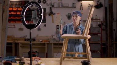 Eski ahşap sandalyeyi öğütme makinesiyle yeniden boyayan yaşlı kadın blogcu eski kaplama ve kayıt videosunu kaldırmak için. blog yazma, mobilya restorasyonu ve ev geliştirme kavramı