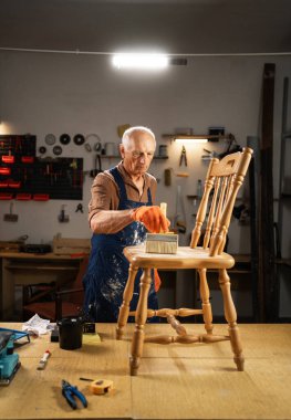 Yaşlı bir adam zımpara kağıdıyla zımparaladıktan sonra ahşap bir sandalyeden tozu temizler. Önlük ve turuncu eldivenli bir işçi atölyesinde çalışır, restorasyon yapar ve bunu kendi başına yapar..