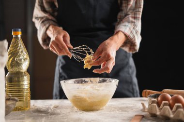 Önlüklü bir fırıncı hamur ve cam kaseyi çırpıcıyla karıştırıyor. Sahne, bir ev mutfağında yağ ve yumurta gibi malzemelerle dikkatlice hazırlanma sürecini yakalıyor..