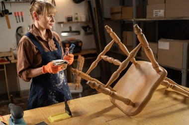 60 yaşında bir kadın elinde çözücü ve bez olan eski bir sandalyeyle bir masanın başında duruyor. Atölyesindeki antika mobilyaları tutkuyla yeniliyor.