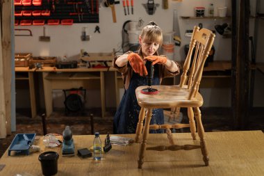 Yaşlı bir kadın atölyede eski bir sandalyeyi tamir ediyor ve bir tahtayı zımparalıyor. Boşluğu kopyala