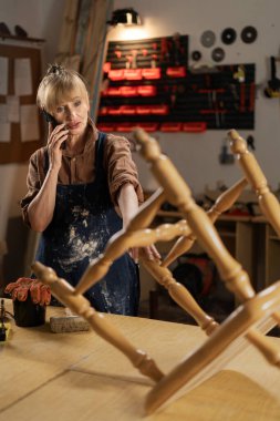 Önlüklü ve gömlekli bir kadın tamirat sırasında telefonla konuşuyor, atölyesinde duruyor. konsept onarımı ve eski mobilyaların restorasyonu.