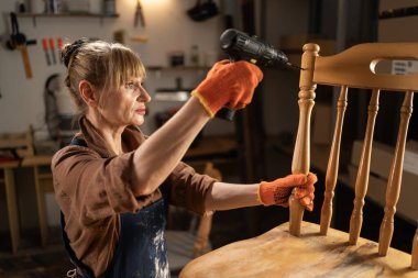 Önlüklü ve gömlekli bir kadın tamirat sırasında sandalyede delikler açıp atölyesinde dikiliyor. konsept onarımı ve eski mobilyaların restorasyonu.