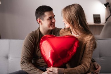 Sevgililer Günün kutlu olsun. Aşık genç bir çift kırmızı bir kalp şeklinde balon tutuyor ve birbirlerine bakıyor oturma odasındaki kanepede oturuyorlar. Birlikte romantik bir akşam. Boşluğu kopyala