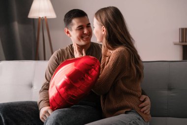 Sevgililer günün kutlu olsun. Kanepede otururken ellerinde kırmızı bir folyo balonu tutan sevimli bir çift. Romantik bir akşam. Boşluğu kopyala