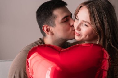 Kanepede oturan, kalp şeklinde balon tutan ve öpüşen bir çiftin portresi. Yaklaş. Sevgililer günün kutlu olsun. Birlikte romantik bir akşam.