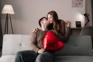Aşık genç bir çift, ellerinde kalp şeklinde folyo balonuyla evde romantik bir akşam geçirir. Sevgilisi kanepede otururken kız arkadaşını yanağından öper. Sevgililer Günün kutlu olsun.