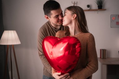 Sevgililer Günün kutlu olsun. Birbirine aşık genç bir çift evdeki oturma odasında kalp şeklinde bir balonu öpüyor ve tutuyorlar. İlişki ve romantik bir akşam. Boşluğu kopyala