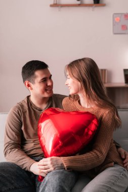 Genç çift oturma odasında kanepede oturup birbirlerine bakıyorlar ve ellerinde kırmızı kalp şeklinde bir balon tutuyorlar. Romantik bir akşam.