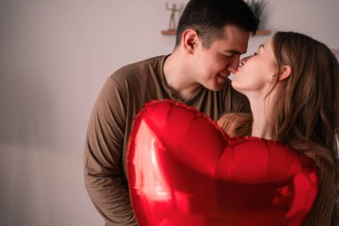 Sevgililer Günün kutlu olsun. Birbirine aşık genç bir çift evdeki oturma odasında kalp şeklinde bir balonu öpüyor ve tutuyorlar. İlişki ve romantik bir akşam. Boşluğu kopyala