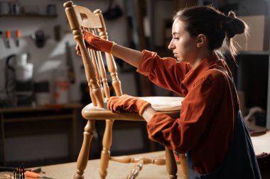 Mobilya yenileme, restorasyon ve loş konsept. Genç ve çekici bir kadın atölyesinde çalışan eski ahşap sandalyeyi zımparalıyor. Boşluğu kopyala