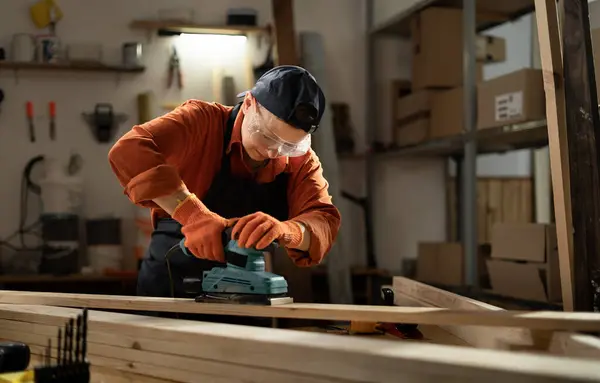 stock image Female carpenter in safety glasses sanding wooden board using belt sander, working in her workshop. Copy space