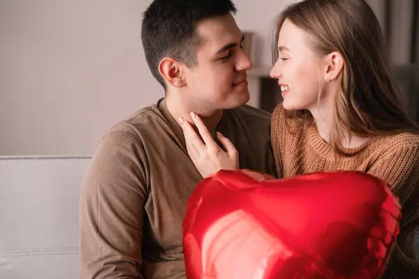 stock image Happy Valentine's Day concept. Lovely couple holding a red heart-shaped foil balloon while sitting on the sofa. Romantic evening. Copy space