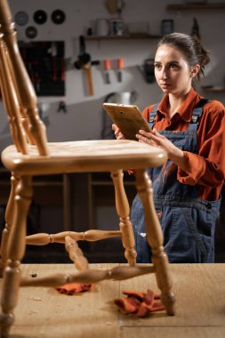 Young woman craftsman trainee with digital tablet standing near wooden table working in the carpentry workshop, copy space clipart