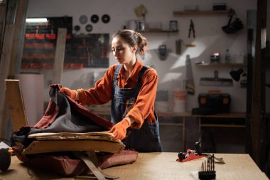 Atölyede çalışan genç bir kadın, yenilenme sürecindeki işçiliğe ve detaylara odaklanmak için koltuğun yerine kumaşı döşeyerek eski mobilyaları restore ediyor. Boşluğu kopyala