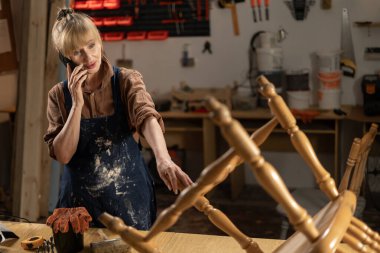 Önlüklü ve gömlekli bir kadın tamirat sırasında telefonla konuşuyor, atölyesinde duruyor. konsept onarımı ve eski mobilyaların restorasyonu.