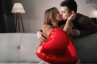 Sevgililer günün kutlu olsun. Genç çift, oturma odasında kanepede otururken öpüşüp, kalp şeklinde bir balon tutuyorlar. Boşluğu kopyala