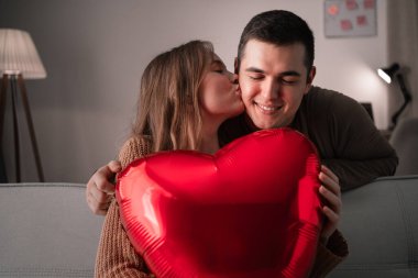 Aşık genç bir çift oturma odasında kalp şeklinde bir balon tutuyor, bir kadın sevgilisini yanağından öpüyor. Sevgililer günün kutlu olsun.