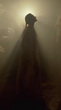 Örgü örmüş bir kadının silüeti, plastik bir çarşaf ve sisle hafifçe aydınlatılmış.