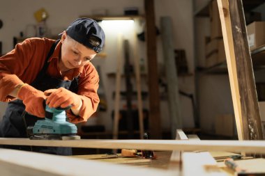 Zanaatkar ve DIY el yapımı. Marangozluk atölyesinde elektrikli zımpara makinesi kullanan kadın marangoz, ahşap kalasları zımparalıyor. Boşluğu kopyala