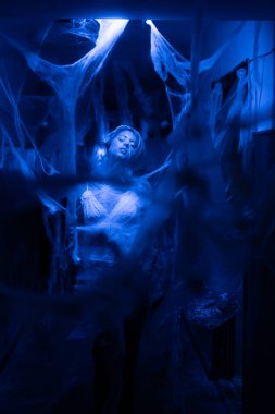 Bir kadın, ağlarla kaplı mavi ışıklı bir koridorda duruyor. Duygusal tuzağa düşmeyi ve gerçeküstü iç gözlemleri sembolize ediyor..