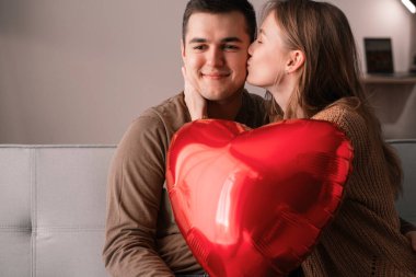 Kanepede oturan genç bir çiftin portresi, ellerinde kırmızı kalp şeklinde balon var. Aşk ve Sevgililer Günü konsepti